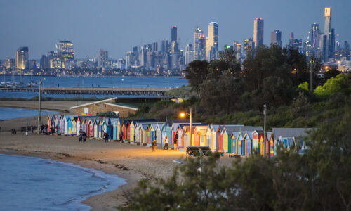 Melbourne, Australia - December 8, 2017: Visitors around beach boxes at Brighton Beach at dusk. Skyscrapers in the city visible in the distance.