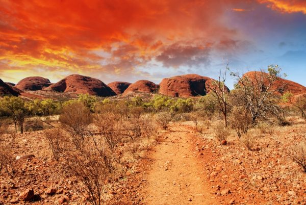 A dirt path leads toward large, red rock formations under an orange and blue sky.