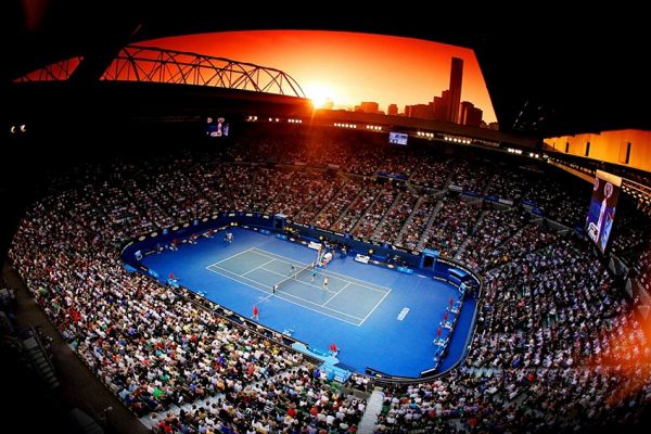 An overhead view of a tennis match in a large stadium at sunset, with a red sky over the city skyline in the background.