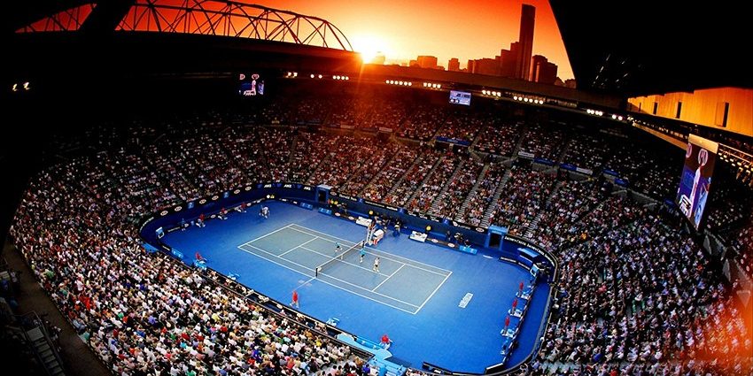 An overhead view of a tennis match in a large stadium at sunset, with a red sky over the city skyline in the background.