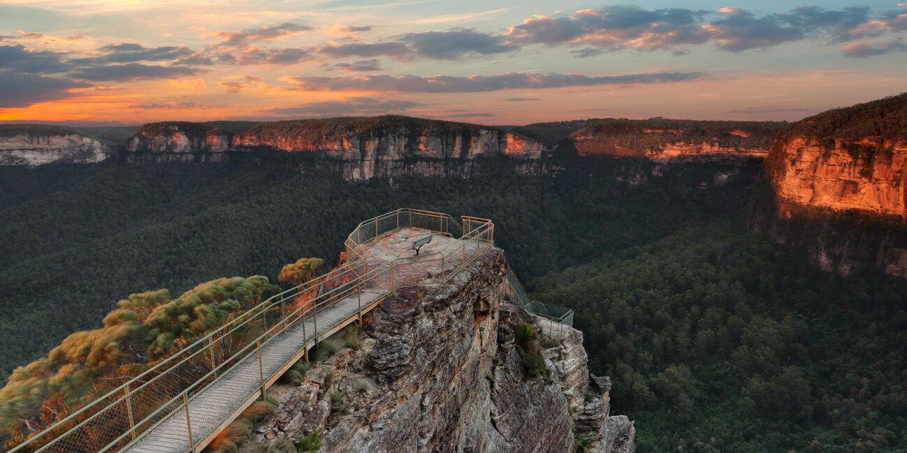 A lookout platform on a cliff overlooking a forested valley in the Blue Mountains at sunset.