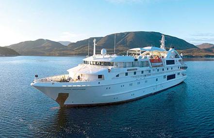 A large white cruise ship named Coral Discoverer floats on a lake with hills in the background.